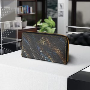 May include: A rectangular zip-around wallet featuring a dark, textured surface with abstract streaks of blue and gold. The centre of the wallet displays an elegant, gold-coloured monogram with the intertwined letters AC in a cursive script font.