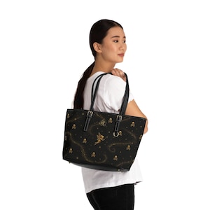 Puede incluir: Un bolso tote negro con un patrón dorado de una pequeña hada alada y motivos de calaveras. El bolso tiene dos correas negras para el hombro con hebillas plateadas y un diseño de fondo oscuro y estrellado con polvo de oro en espiral.