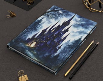 May include: A hardcover journal featuring a dark blue castle illustration set in a forest. The cover art depicts a gothic castle with tall spires, surrounded by trees under a cloudy sky. Gold and black pencils and gold binder clips are also visible.