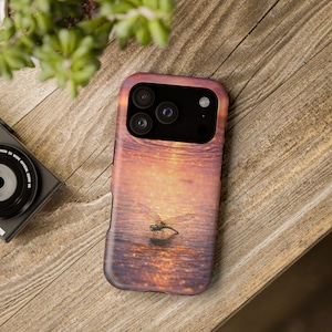 Puede incluir: Funda de teléfono con una escena de puesta de sol y una libélula en el agua. La funda tiene un diseño envolvente con tonos naranja, amarillo y morado. La funda del teléfono está sobre una superficie de madera.
