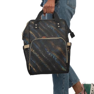 Puede incluir: Una mochila negra con un patrón texturizado de piel de serpiente iridiscente con rayas diagonales azules y doradas. La bolsa incluye dos asas superiores, un bolsillo frontal con cremallera y borde beige, bolsillos laterales y cremalleras de metal dorado.