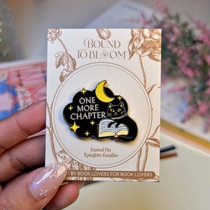 May include: An enamel pin featuring a black cat reading a book under a yellow crescent moon and stars, set against a black cloud. The pin reads "ONE MORE CHAPTER" and is on a card with floral designs and the text "BOUND TO BLOOM".