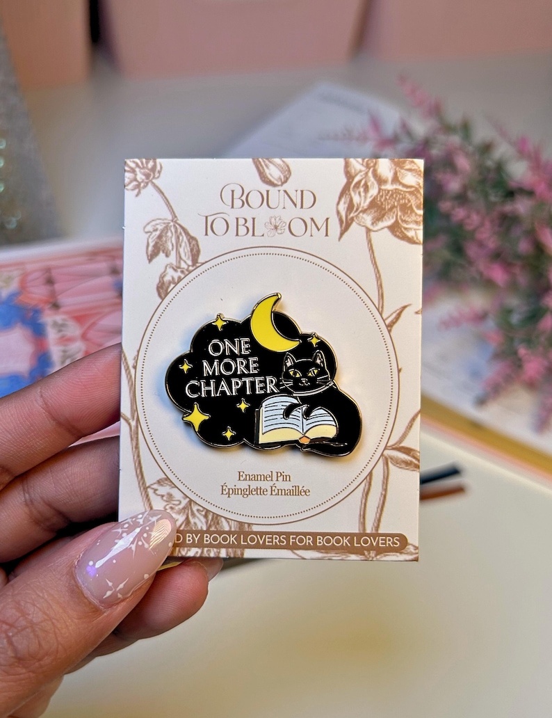 May include: An enamel pin featuring a black cat reading a book under a yellow crescent moon and stars, set against a black cloud. The pin reads "ONE MORE CHAPTER" and is on a card with floral designs and the text "BOUND TO BLOOM".