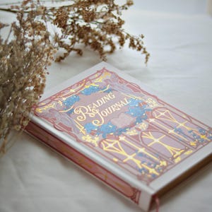 Könnte beinhalten: Ein Hardcover "Reading Journal" mit rosa Einband und goldener Schrift. Das Cover zeigt ein verziertes Design mit blauen floralen Akzenten und einem rosa Lesezeichen. Das Journal befindet sich vor einem weißen Hintergrund mit getrockneten Blumen.