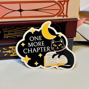 May include: A black sticker with the text "ONE MORE CHAPTER" and a cartoon cat reading a book under a yellow crescent moon and stars. The cat wears a pink collar. The sticker is on a white surface with books in the background.