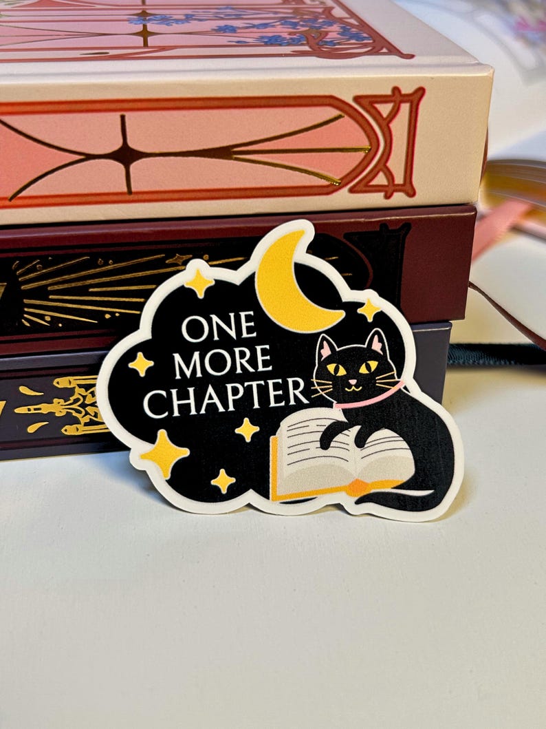 May include: A black sticker with the text "ONE MORE CHAPTER" and a cartoon cat reading a book under a yellow crescent moon and stars. The cat wears a pink collar. The sticker is on a white surface with books in the background.