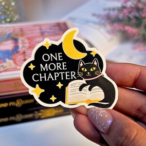 May include: A black sticker with a cat reading a book, a yellow crescent moon, and the words "ONE MORE CHAPTER". The cat has yellow eyes and a pink collar. The sticker is surrounded by yellow stars and a black cloud.