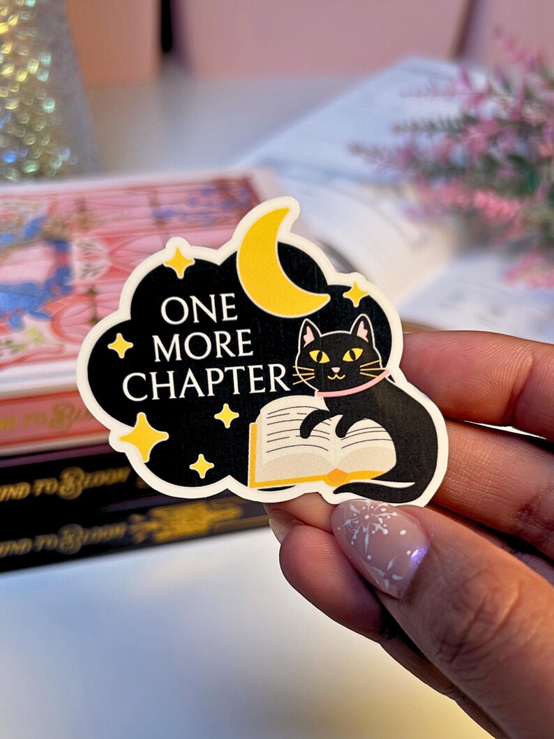 May include: A black sticker with a cat reading a book, a yellow crescent moon, and the words "ONE MORE CHAPTER". The cat has yellow eyes and a pink collar. The sticker is surrounded by yellow stars and a black cloud.