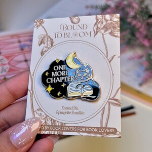 May include: Enamel pin featuring a black cat reading a book under a crescent moon and stars. The pin is set against a black cloud with the words "ONE MORE CHAPTER". The pin is on a card that says "BOUND TO BLOOM" and "ED BY BOOK LOVERS FOR BOOK LOVERS".