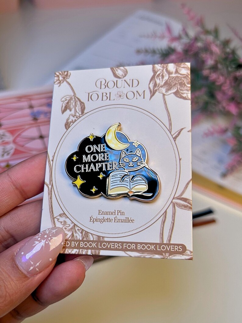 May include: Enamel pin featuring a black cat reading a book under a crescent moon and stars. The pin is set against a black cloud with the words "ONE MORE CHAPTER". The pin is on a card that says "BOUND TO BLOOM" and "ED BY BOOK LOVERS FOR BOOK LOVERS".