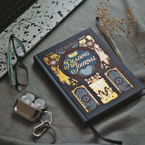 May include: A Reading Journal with gold accents and the title in elegant script sits on a gray surface, next to a laptop, eyeglasses, and wireless earbuds. A pen and dried flowers are also present, creating a cozy reading nook scene.