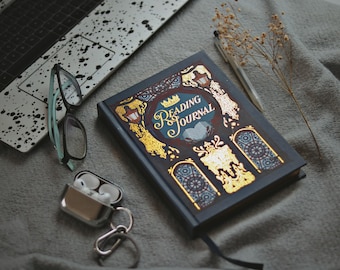 May include: A Reading Journal with gold accents and the title in elegant script sits on a gray surface, next to a laptop, eyeglasses, and wireless earbuds. A pen and dried flowers are also present, creating a cozy reading nook scene.
