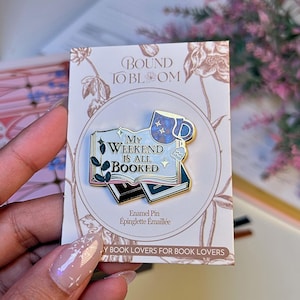 May include: Enamel pin featuring the text "My Weekend Is All Booked" on a stack of books, with a blue mug and floral details. The pin is on a card with the text "Bound to Bloom" and "By Book Lovers for Book Lovers".