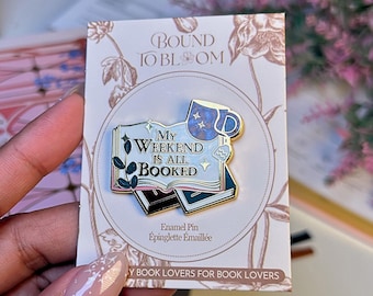 May include: Enamel pin with the text "My Weekend Is All Booked" on a stack of books, with a blue mug and floral accents. The pin is on a card with the text "Bound to Bloom" and "By Book Lovers for Book Lovers".
