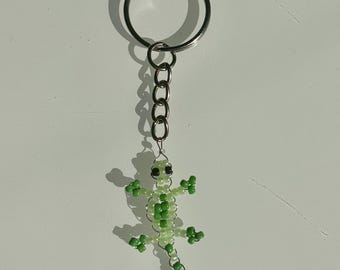 May include: A beaded lizard keychain in shades of green and white. The lizard is crafted with small beads, featuring a detailed body and limbs. The keychain includes a silver-colored ring and chain.