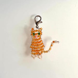 May include: A beaded cat charm with orange and white beads. The cat has green eyes and a pink nose. The charm is attached to a silver-colored clasp. The cat's tail is curved.
