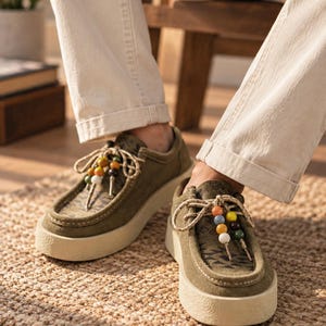 May include: Olive green suede shoes with a thick, textured off-white rubber sole. The upper features a dark geometric pattern on the tongue and toe box. The beige laces are adorned with a row of small, colorful beads in yellow, blue, and white.