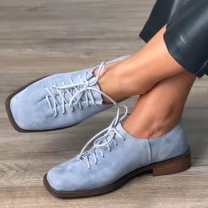 May include: Pale blue suede oxford shoes with square toes and brown soles. The shoes feature white laces and a low heel. These shoes are a stylish choice for casual or semi-formal wear.