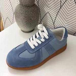 May include: A blue suede trainer with white laces and a brown sole. The shoe has a retro design with a padded tongue and collar. The trainer is a casual style, suitable for everyday wear.