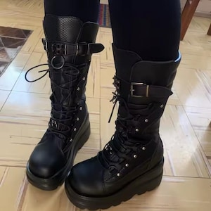 May include: Pair of black platform boots with multiple buckles, laces, and a textured upper. The boots have a chunky sole and a tall shaft, with a mix of smooth and textured leather. The boots are designed with a gothic or punk aesthetic.