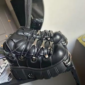 Puede incluir: Botas de plataforma negras con detalles metálicos plateados. Las botas presentan un diseño con cordones con cierres metálicos decorativos y una suela gruesa y texturizada. Fabricadas con un material liso similar al cuero.