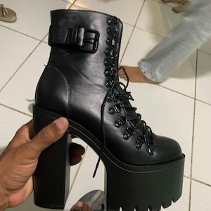 Könnte beinhalten: Schwarzer Plateau-Stiefel mit klobigem Absatz und Schnürdesign. Der Stiefel hat einen Schnallenverschluss am Knöchel und eine dicke, profilierte Sohle. Der Absatz des Stiefels misst ca. 13 cm.