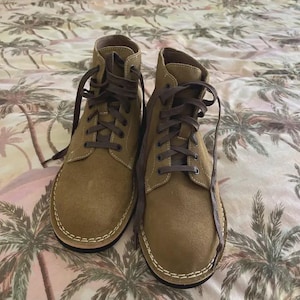 May include: A pair of tan suede ankle boots featuring dark brown laces, a rounded toe, and prominent white contrast stitching along the sole edge. The boots have a high-top design with metal eyelets and a flat black outsole for a sturdy appearance.