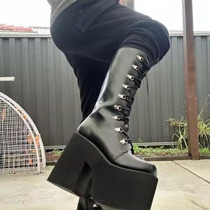 Könnte beinhalten: Schwarze Plateau-Stiefel mit Schnürung und silberfarbenen Ösen. Die Stiefel haben einen hohen Schaft und eine dicke, rechteckige Plateausohle. Die Stiefel sind aus glänzendem Material gefertigt.