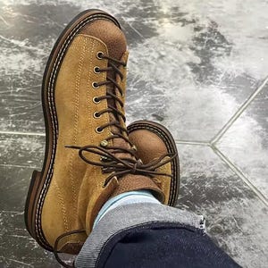 May include: A tan suede lace-up boot featuring a textured brown leather tongue and collar, dark brown laces, brass eyelets, and a thick dark brown sole with visible white stitching along the welt, worn with dark blue denim jeans and light blue socks.