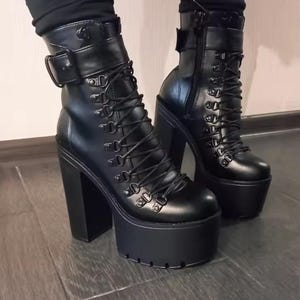 May include: A pair of black ankle boots featuring a high block heel and a thick platform sole with a lugged tread. The boots have a lace-up front with metal eyelets, a side zip closure, and an adjustable strap with a buckle at the top collar.