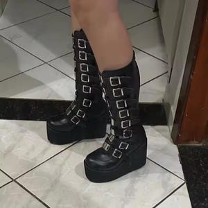 May include: Black knee-high platform boots with multiple silver buckles. The boots have a thick platform sole and a wedge heel. The leather boots are a bold fashion statement.