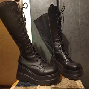 May include: A pair of black knee-high boots featuring a thick platform sole with a scalloped edge, a rounded toe, and a full-length lace-up front closure with black laces. The boots have a side zipper and a smooth, matte finish throughout the exterior.