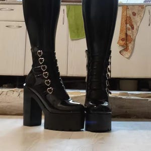 Può includere: Paio di stivali neri con plateau e finitura lucida. Gli stivali hanno un tacco grosso, una suola platform e dettagli a forma di cuore. Uno stivale ha una cerniera, l'altro fibbie a forma di cuore. Gli stivali sono al ginocchio.