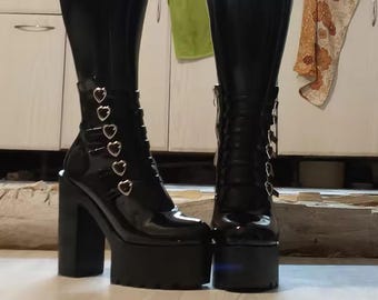 Puede incluir: Par de botas negras de plataforma con acabado brillante. Las botas tienen tacón grueso, suela de plataforma y detalles en forma de corazón. Una bota tiene cremallera, la otra hebillas en forma de corazón. Las botas son hasta la rodilla.