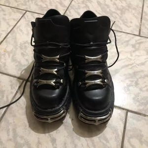 May include: A pair of black leather ankle boots featuring a thick platform sole, black laces, and three silver-toned metal plates across the front of each boot. The toe area of each boot is reinforced with a silver-toned metal cage-like detail.