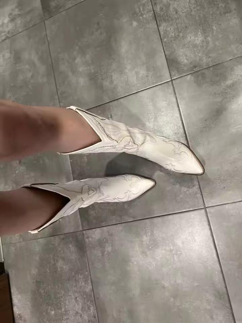 Op de afbeelding: Een paar witte leren cowboylaarzen met een puntige neus en gedetailleerde ton-sur-ton stikselpatronen op de wreef en de schacht. De laarzen hebben een halflange schacht met een V-vormige opening aan de bovenkant en een blokhak.