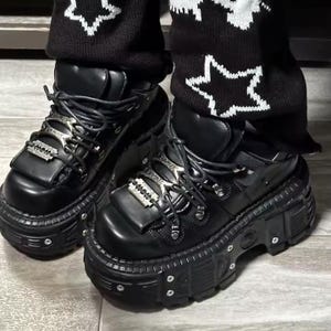 May include: Black platform boots with silver-tone accents. The boots feature a chunky sole, lace-up closure, and decorative metal details. The boots are worn with black pants with white star patterns.