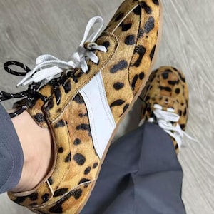 May include: Leopard print trainers with white laces and a white side stripe. The shoes have a brown sole and black and white laces. These trainers are a fashionable footwear choice.