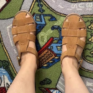 May include: A pair of tan suede sandals with a woven design. The sandals feature an ankle strap and buckle fastening. The shoes are on a patterned rug.