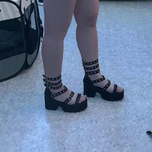 May include: Black platform sandals with multiple straps and silver-coloured studs. The sandals have a chunky heel and are worn on a light-coloured surface. The straps wrap around the ankle and foot.