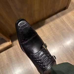 May include: A black shoe featuring a square toe and a textured snakeskin pattern on the toe cap and strap area. The shoe includes two metallic buckles on the side, securing the strap across the top of the foot, with a smooth finish on the heel.