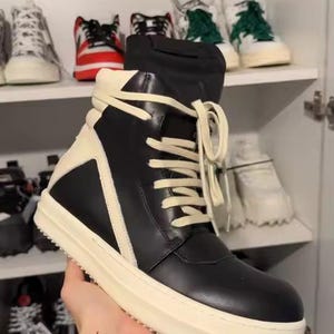 May include: A high-top sneaker featuring a black leather upper with cream-colored leather overlays and a matching cream-colored thick rubber sole. The shoe includes cream-colored laces and a black fabric tongue extending above the collar.