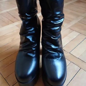Könnte beinhalten: Ein Paar schwarze wadenhohe Stiefel mit einer glatten, halbglänzenden Oberfläche und einem markant gerafften Schaftdesign. Diese Stiefel sind mit einer dicken, massiven Plateausohle und einer abgerundeten Zehenpartie konstruiert.