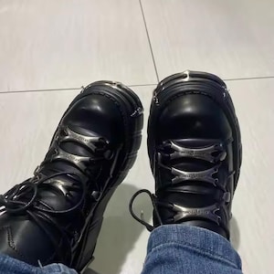 May include: A pair of black leather boots featuring thick soles, rounded toes with silver-toned metal caps, and black laces threaded through three horizontal silver-toned metal plates on the front of each boot, worn with blue denim jeans.