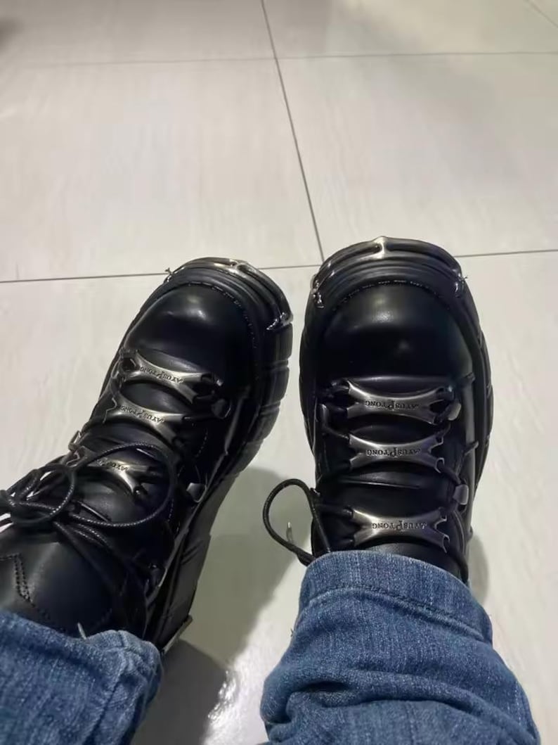 May include: A pair of black leather boots featuring thick soles, rounded toes with silver-toned metal caps, and black laces threaded through three horizontal silver-toned metal plates on the front of each boot, worn with blue denim jeans.