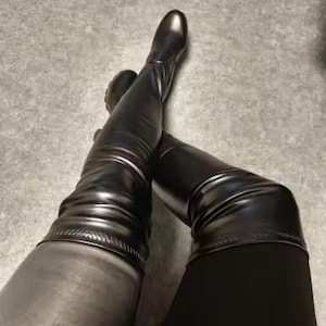 May include: A pair of black over-the-knee boots featuring a smooth, semi-gloss finish. The boots have a rounded toe, a fitted shaft that extends above the knee, and a visible seam detail along the upper edge. The material appears flexible and form-fitting.