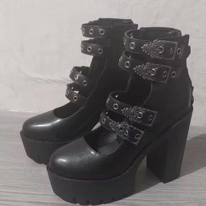 May include: Black platform boots with multiple straps and silver-tone buckles. The boots have a chunky heel and a thick platform sole. The straps are secured with metal eyelets. The style is reminiscent of gothic or alternative fashion.