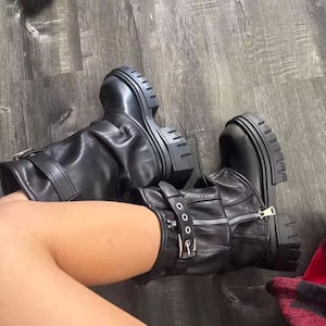 May include: A pair of black leather boots with a chunky, lug sole. The boots have a mid-calf height, with a zipper and buckle details. The boots are shown from a top-down perspective.