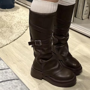 May include: A pair of dark brown mid-calf boots featuring a slouchy shaft design, a wrap-around strap with a silver-toned metal buckle, and a thick, chunky platform sole. The boots are worn with white ribbed socks that extend above the boot tops.