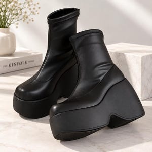 Pode incluir: Um par de botins pretos com acabamento mate liso, biqueira arredondada e sola de plataforma alta com cunha. As botas têm um cano justo que se estende acima do tornozelo com uma costura vertical ao longo do centro da parte frontal.